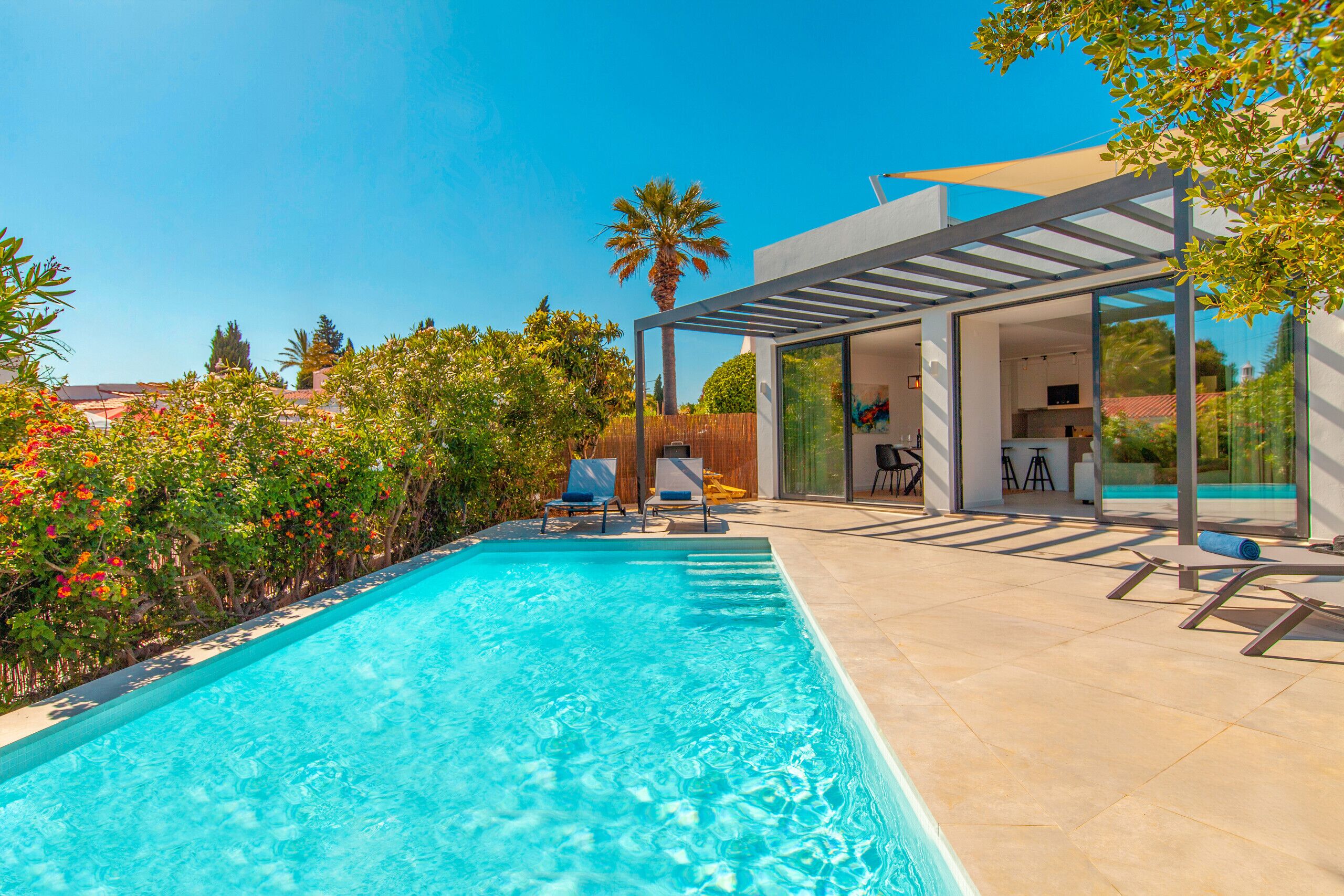 Ein Pool mit Liegestühlen und einer Pergola, umgeben von üppiger Bepflanzung und einem modernen Haus.