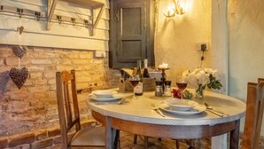 Un coin repas rustique avec une table ronde dressée pour deux personnes, agrémenté d’une nappe blanche à pois, de chaises en bois et d’un mur en briques en arrière-plan.