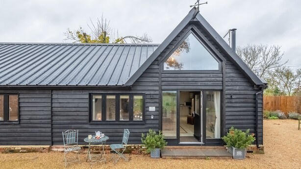 Un edificio de madera negra con techo triangular, un pequeño patio con mesa y sillas, y un jardín con plantas en macetas.