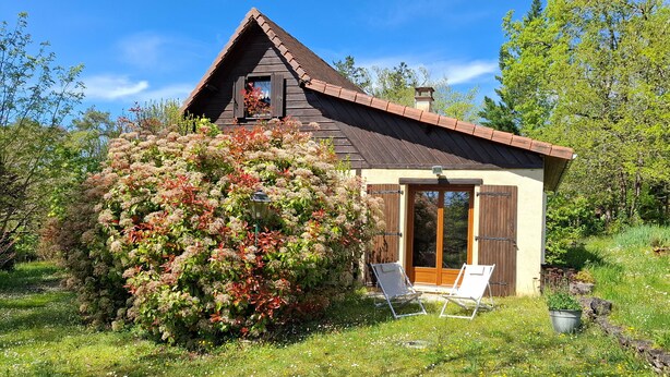 A small wooden cabin with a sloped roof, a large window, and a door with wooden shutters. There are two white lounge chairs outside.