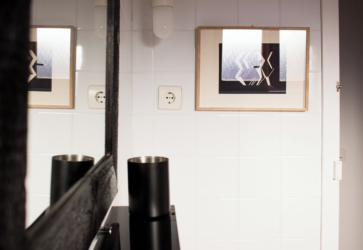 Une salle de bain avec des murs recouverts de carreaux blancs, un lavabo noir et des œuvres d’art encadrées accrochées au mur.
