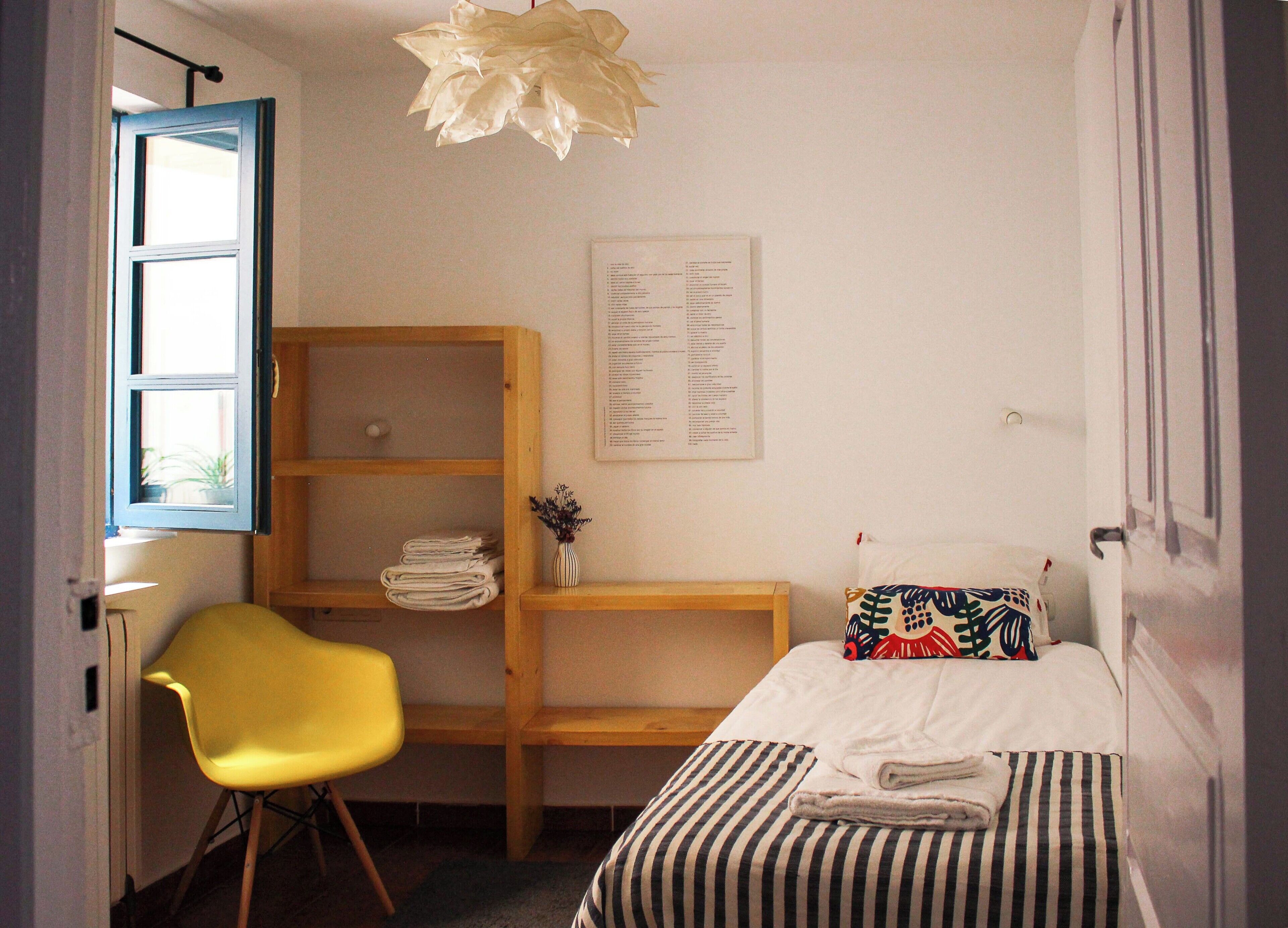 Un dormitorio con una cama, un sillón amarillo, un escritorio de madera con estantes y una lámpara colgante.