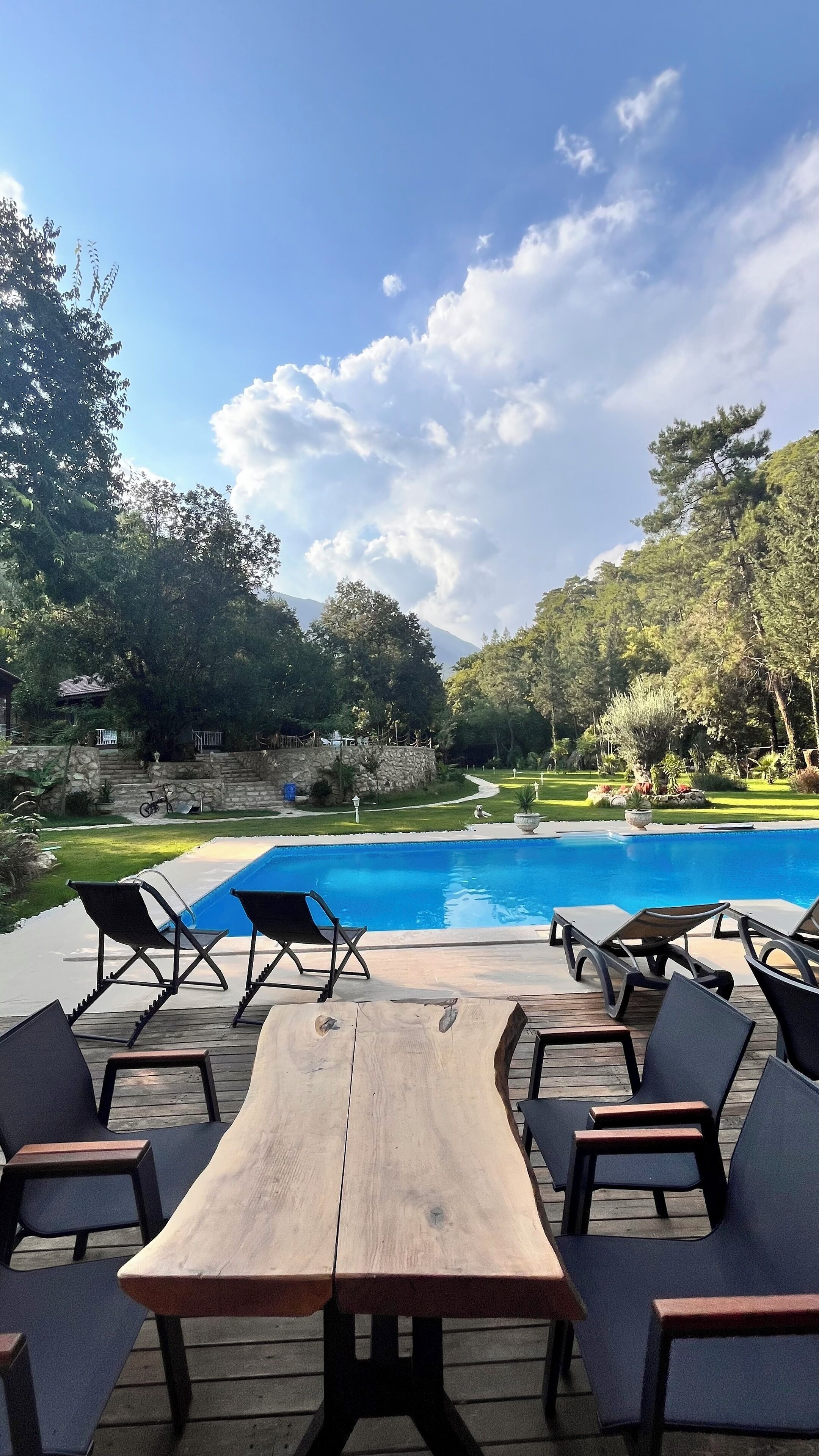 A poolside area with lounge chairs and a wooden table, surrounded by greenery and a stone wall.