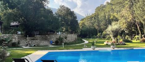 A poolside area with lounge chairs and a wooden table, surrounded by greenery and a stone wall.