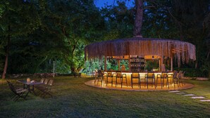 An outdoor bar area with a thatched roof, surrounded by chairs and tables, set in a lush garden with trees. - YUSALİS GARDEN HOTEL (Kumluca)