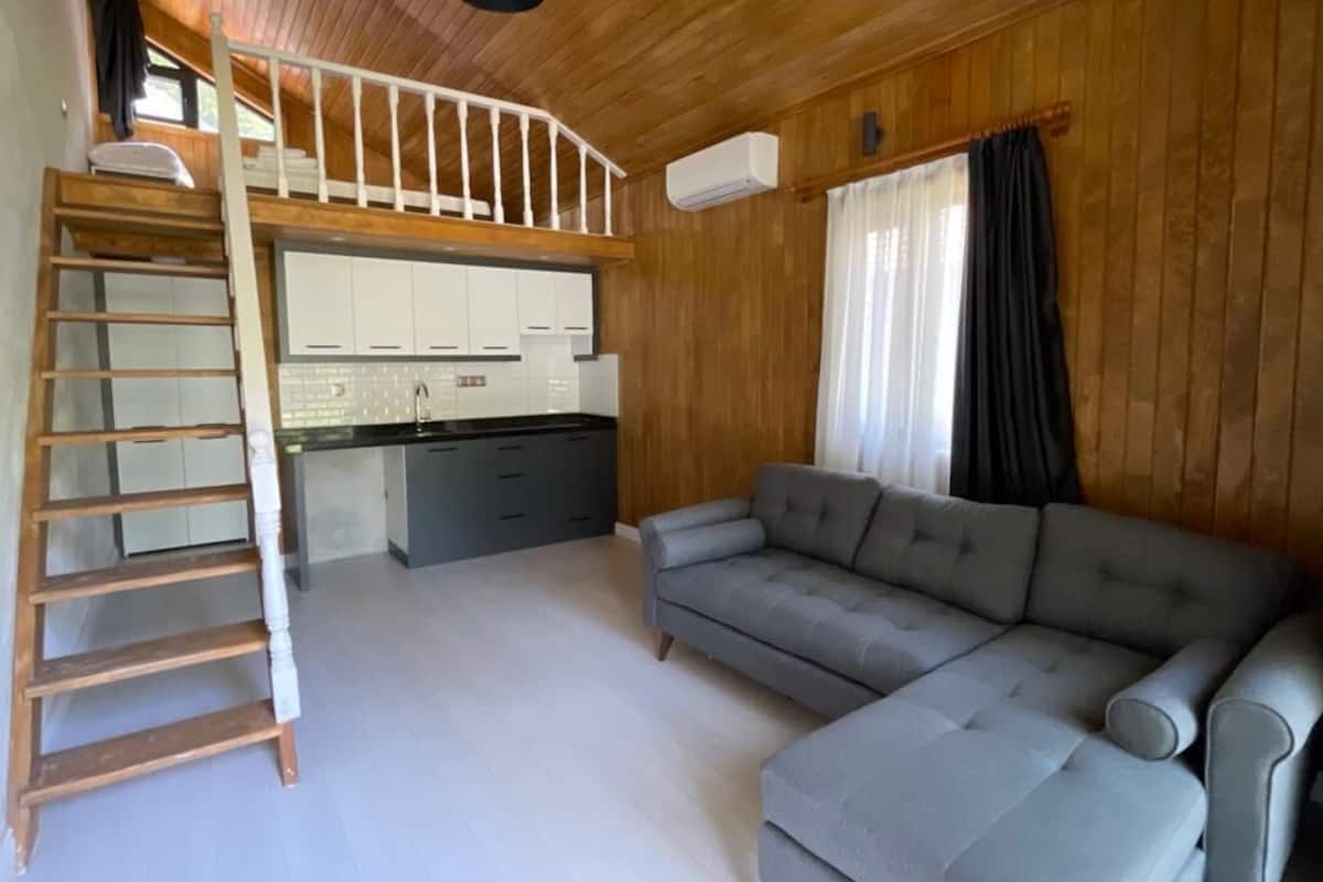 A loft-style room with a wooden ceiling, a gray sofa, a wooden ladder, and a kitchen area with white cabinets.
