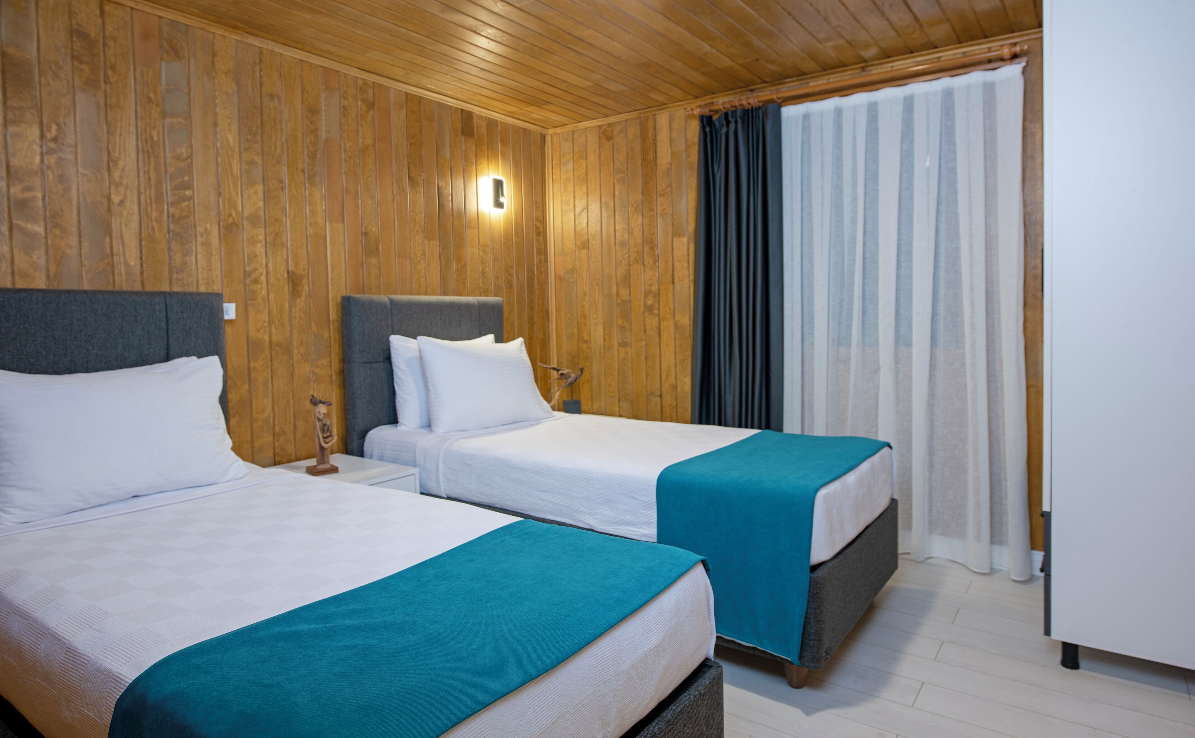 Deux lits simples avec des têtes de lit, un mur en bois, une table de chevet avec une lampe et une fenêtre avec des rideaux.