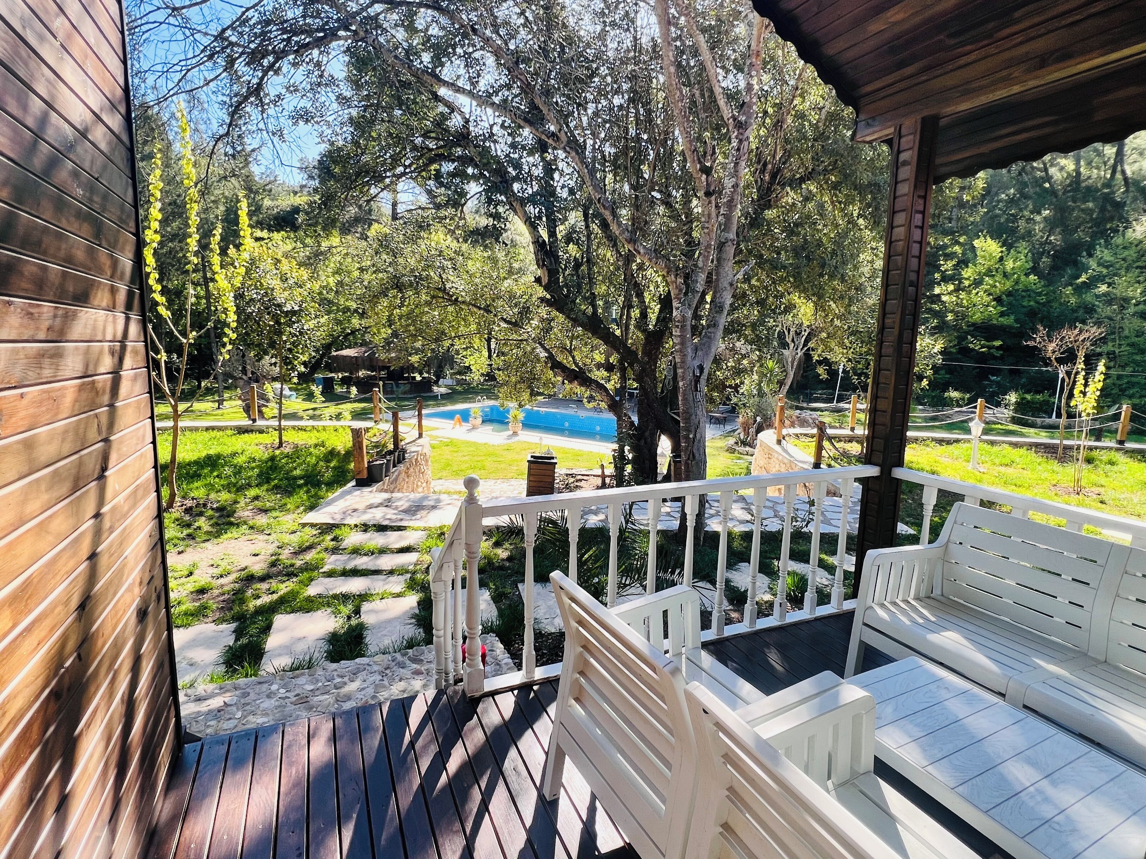 A wooden deck with white railings, a pool, and a garden with trees and a gazebo.