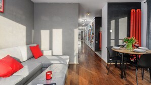 Uma sala de estar moderna com um sofá cinzento, almofadas vermelhas e uma mesa de centro. É visível, ao fundo, uma área de refeições com uma mesa e cadeiras de madeira.