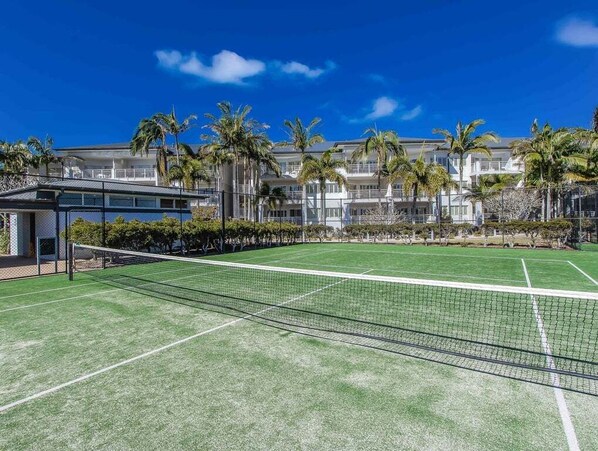 A tennis court with a net, surrounded by palm trees and a building with balconies.