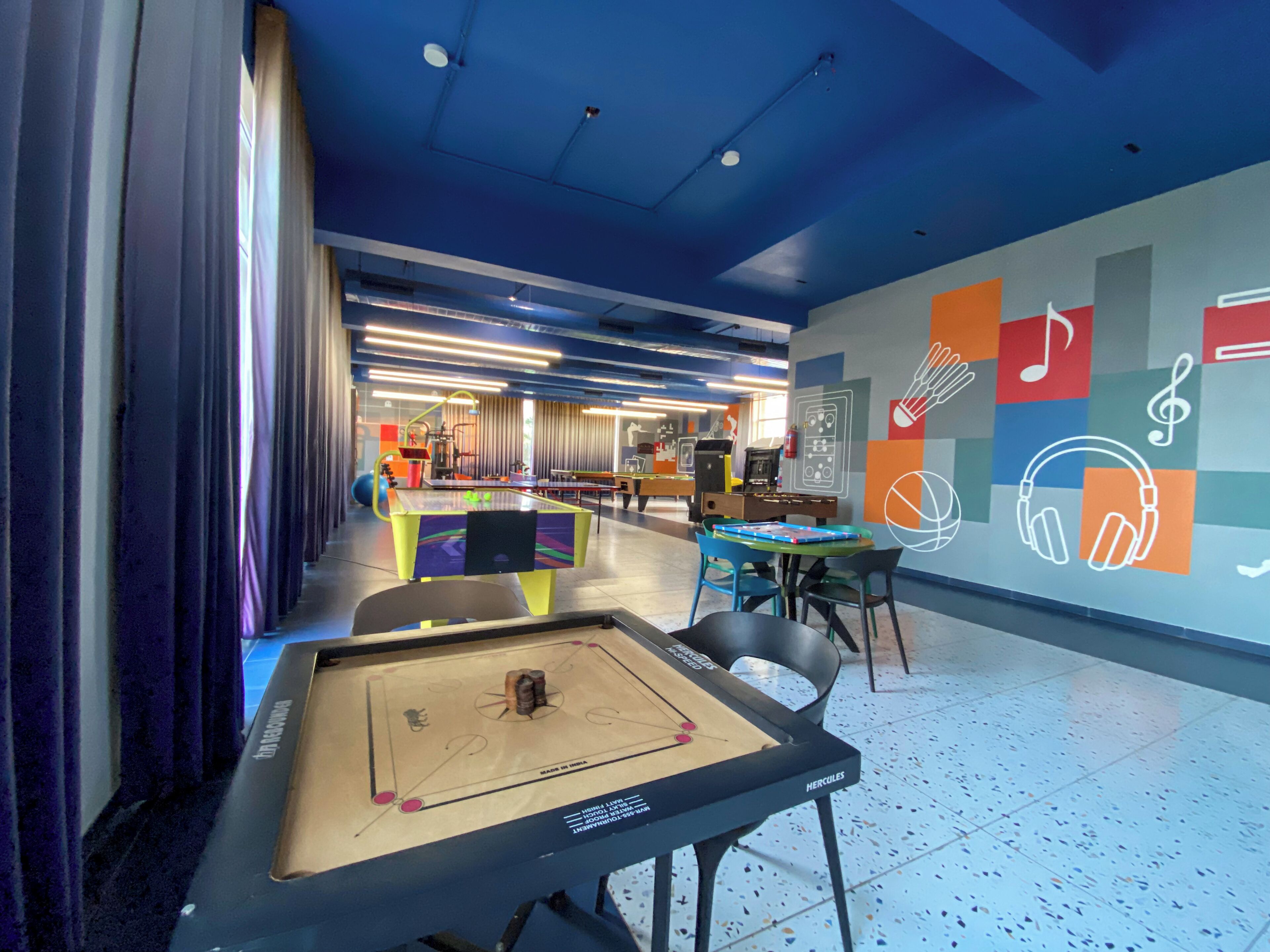 Een moderne ruimte met een tafelvoetbaltafel, een pooltafel en een pingpongtafel. Er zijn kleurrijke wanddecoraties en een plafond met blauwe verlichting.
