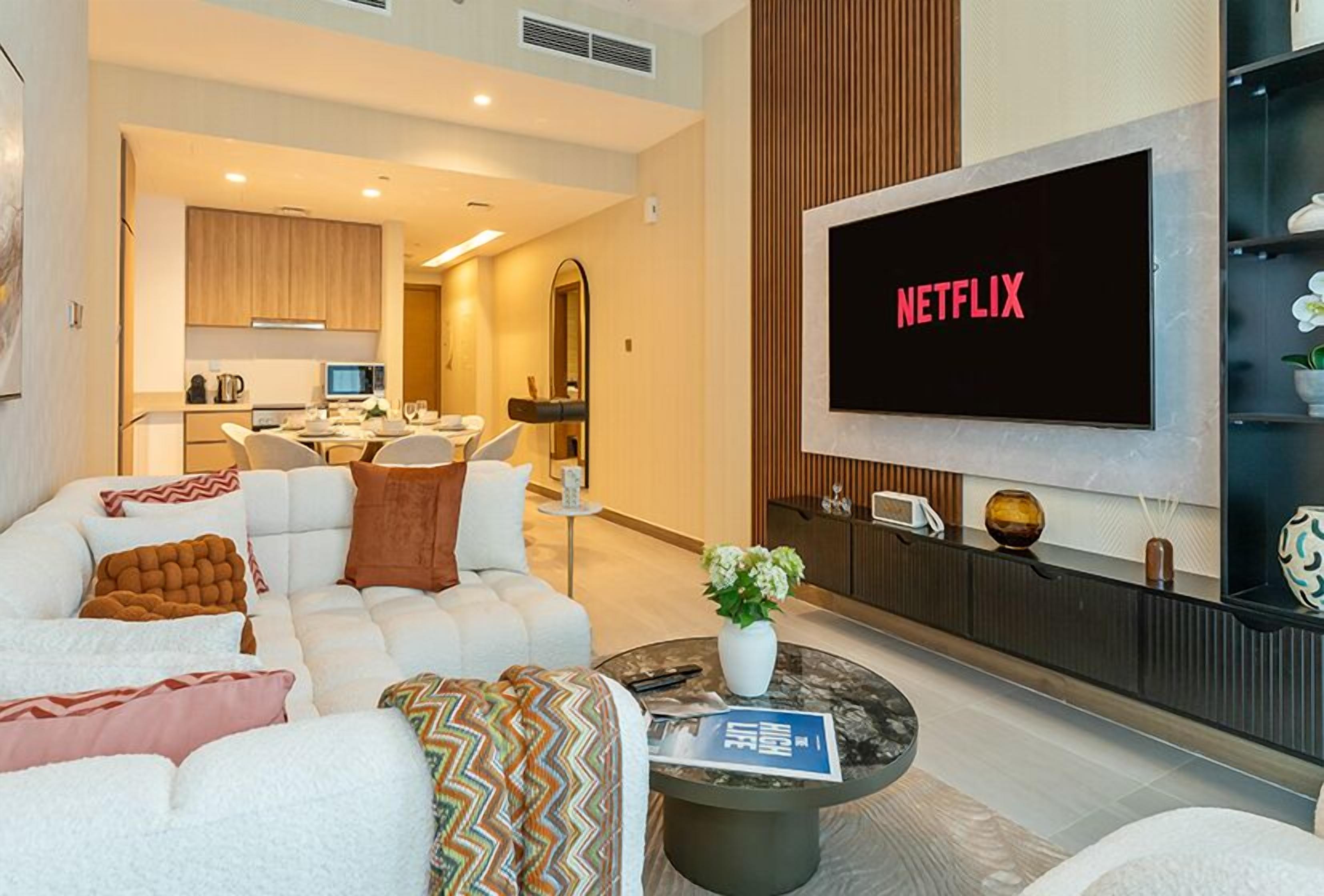 A modern living room with a large flat-screen TV displaying "NETFLIX," a sofa set, a coffee table with a magazine, and a kitchen area in the background.