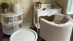 A cozy room with a stone wall, a white armchair, a small round table, and a TV mounted on the wall. - Parvis Cathédrale Bayeux charming studio apartment (Bayeux)