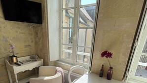 A small, sunlit room with a white table, a white chair, a small white sofa, and a large window. - Parvis Cathédrale Bayeux charming studio apartment (Bayeux)