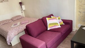A room with a bed and a sofa. The bed has a patterned bedspread and pillows. The sofa has colorful pillows. There is a lamp on a nightstand. - Parvis Cathédrale Bayeux charming studio apartment (Bayeux)