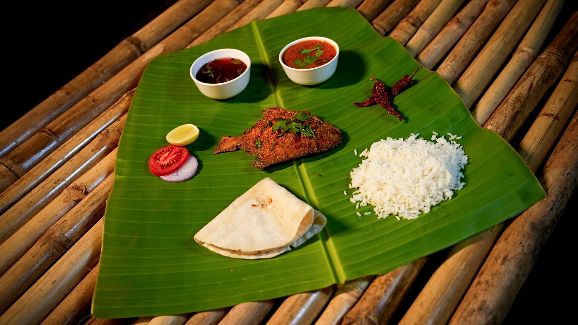 A meal served on a banana leaf with rice, fish, tomato, onion, lemon, and two types of sauce.