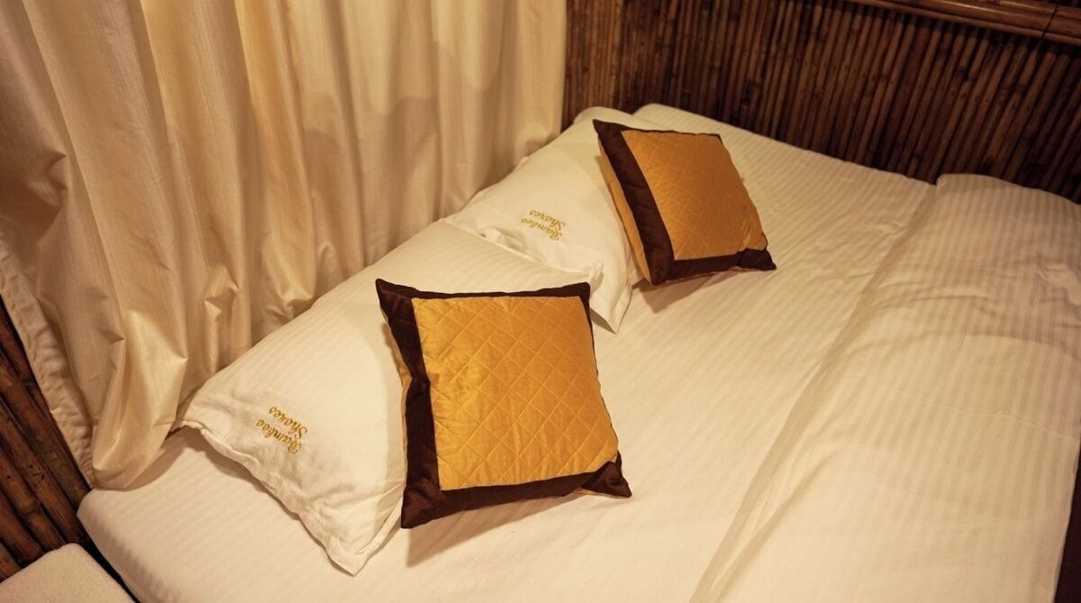 A bed with white linens and two pillows, one with a gold quilted pattern and the other with a brown trim.
