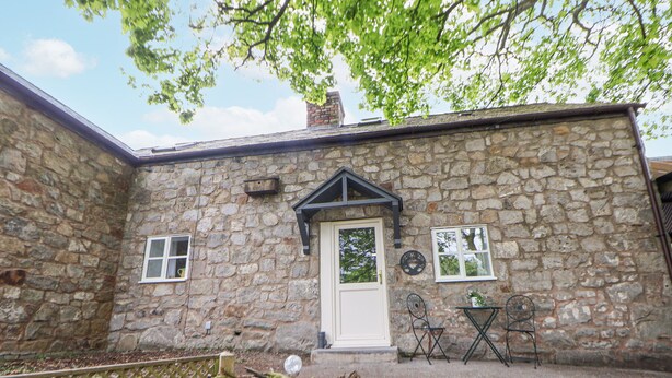 Ein Steinhaus mit einer weißen Tür, zwei Fenstern und einem kleinen Gartenbereich mit Steinen und Pflanzen.