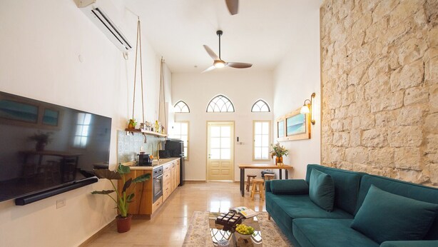 Un salon moderne avec un canapé de couleur turquoise, un téléviseur à écran plat, une cuisine avec un coin repas et un mur en pierre.