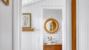 Ein Badezimmer mit weißen Wandverkleidungen, einem Holzschrank, einem Spiegel mit Korbwickerrahmen und einem botanischen Kunstdruck an der Wand.