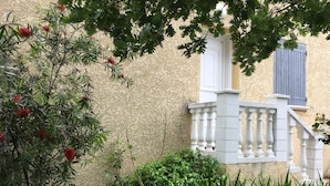 Un bâtiment avec un mur beige à la texture particulière, une porte blanche et un volet bleu. On aperçoit une balustrade blanche sur un escalier menant à une porte.