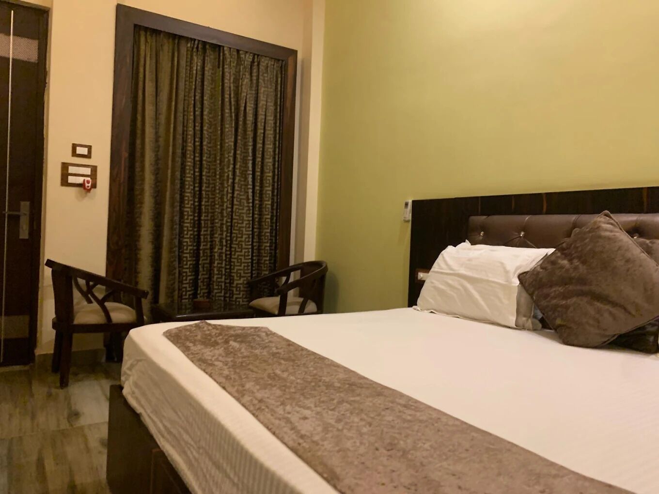 A hotel room with a bed, two chairs, a curtain, and a wall-mounted light switch.
