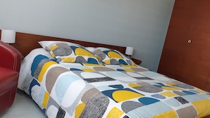 A bed with a colorful geometric-patterned bedspread, a wooden headboard, and a red chair to the left.