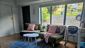 A cozy living room with a sofa and a small table, a blue rug, and a view of the street outside. - Relax and enjoy the North Shore in this cozy and stylish unit. (Auckland)
