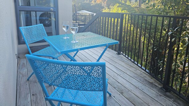 A balcony with a blue metal table and chairs, two wine glasses, and a view of trees and hills. - Relax and enjoy the North Shore in this cozy and stylish unit. (Auckland)