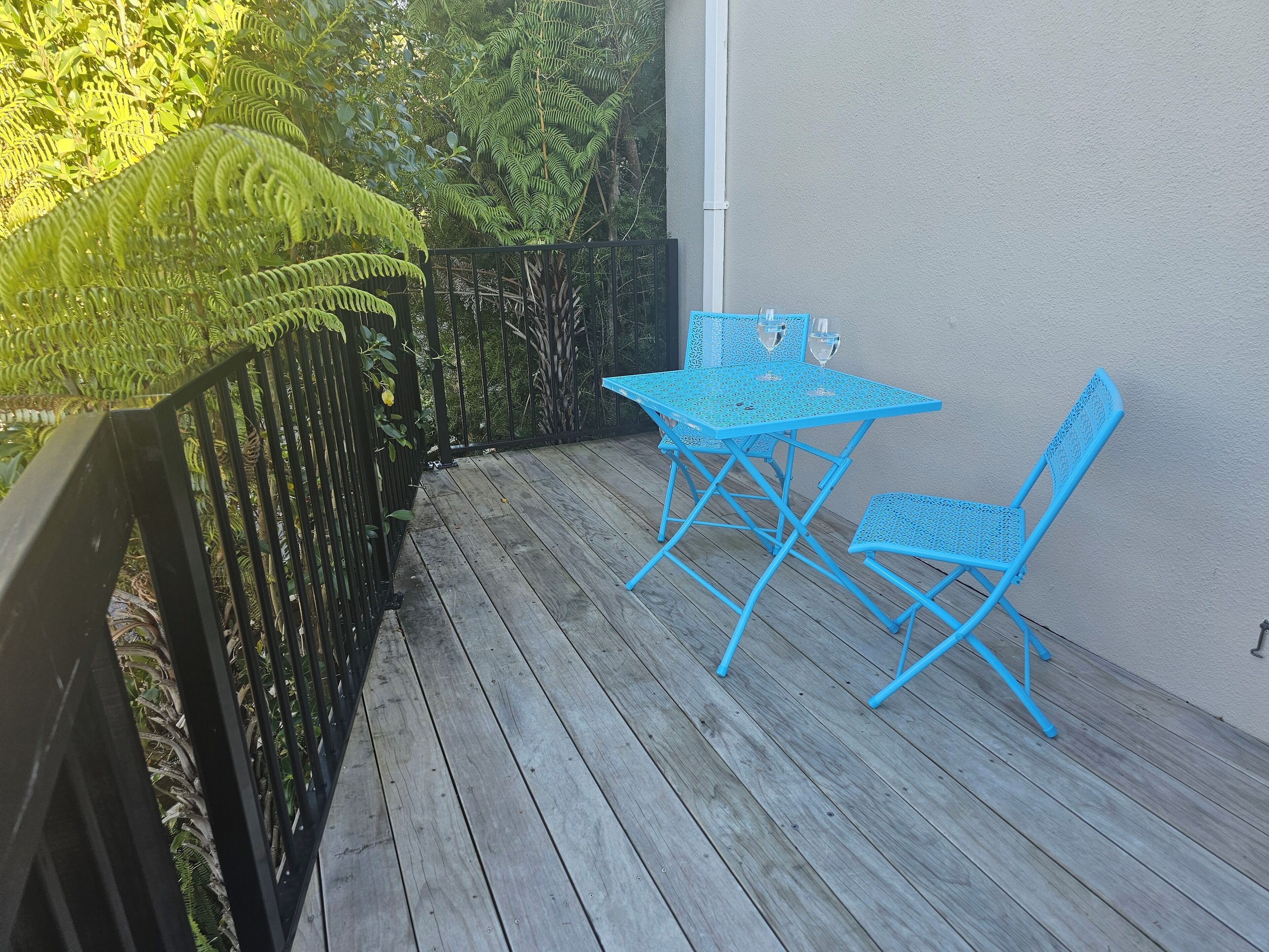 A wooden deck with a blue metal table and two chairs, surrounded by greenery and a black metal fence.