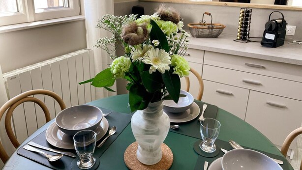 Une table à manger dressée avec des assiettes, des bols et des verres, un vase rempli de fleurs et une fenêtre avec des rideaux.