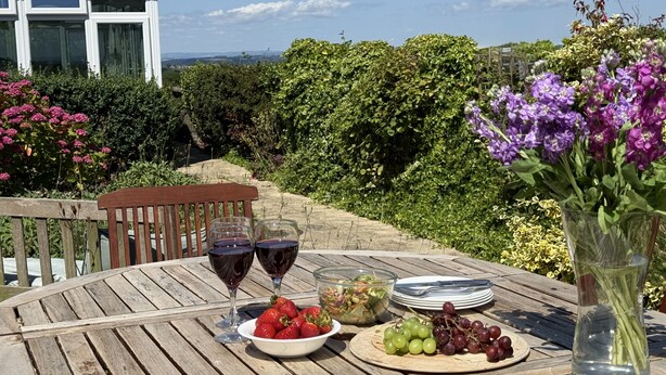 A wooden table set with wine glasses, strawberries, grapes, a salad bowl, and a vase of flowers, overlooking a lush garden and a white building. - Betty's Barn -  Evershot, Dorset (Evershot)
