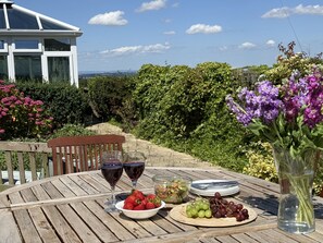Ein Holztisch ist mit Weingläsern, Erdbeeren, Trauben, einer Salatschüssel und einer Blumenvase gedeckt, mit Blick auf einen üppigen Garten und ein weißes Gebäude.