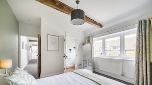 A bedroom with a large bed, a window with curtains, a radiator, and a wooden ceiling beam. - Betty's Barn -  Evershot, Dorset (Evershot)