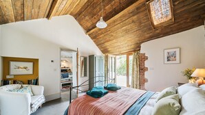 Ein Schlafzimmer mit Holzboden, einem Bett mit vielen Kissen, einem Nachttisch mit Lampe und einem Fenster mit Vorhängen.