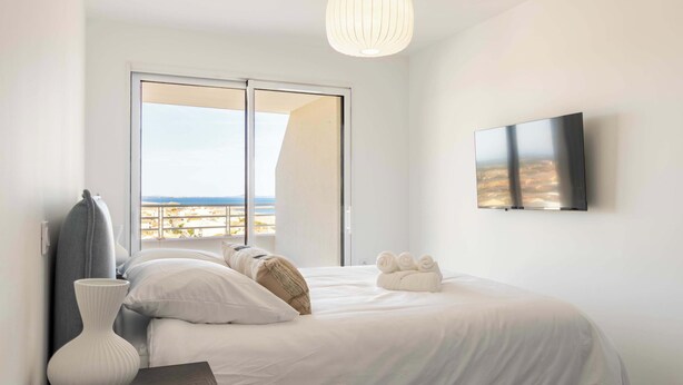 A modern bedroom with a large bed, a view of the beach, a flat-screen TV, and a white vase on a nightstand. - ValrasLoc Panarea with Large Terrace (Valras-Plage)