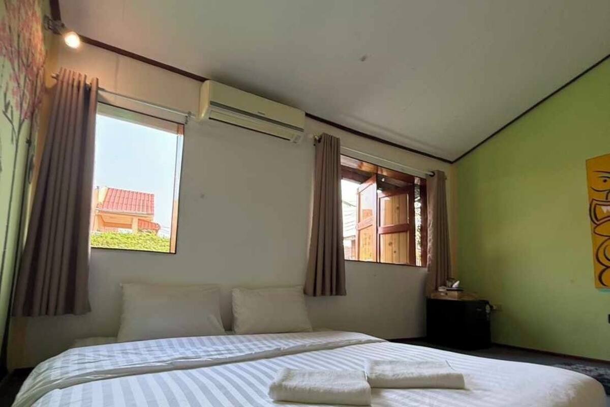 A hotel room with a large bed, two windows with curtains, an air conditioning unit, and a small table with a lamp.