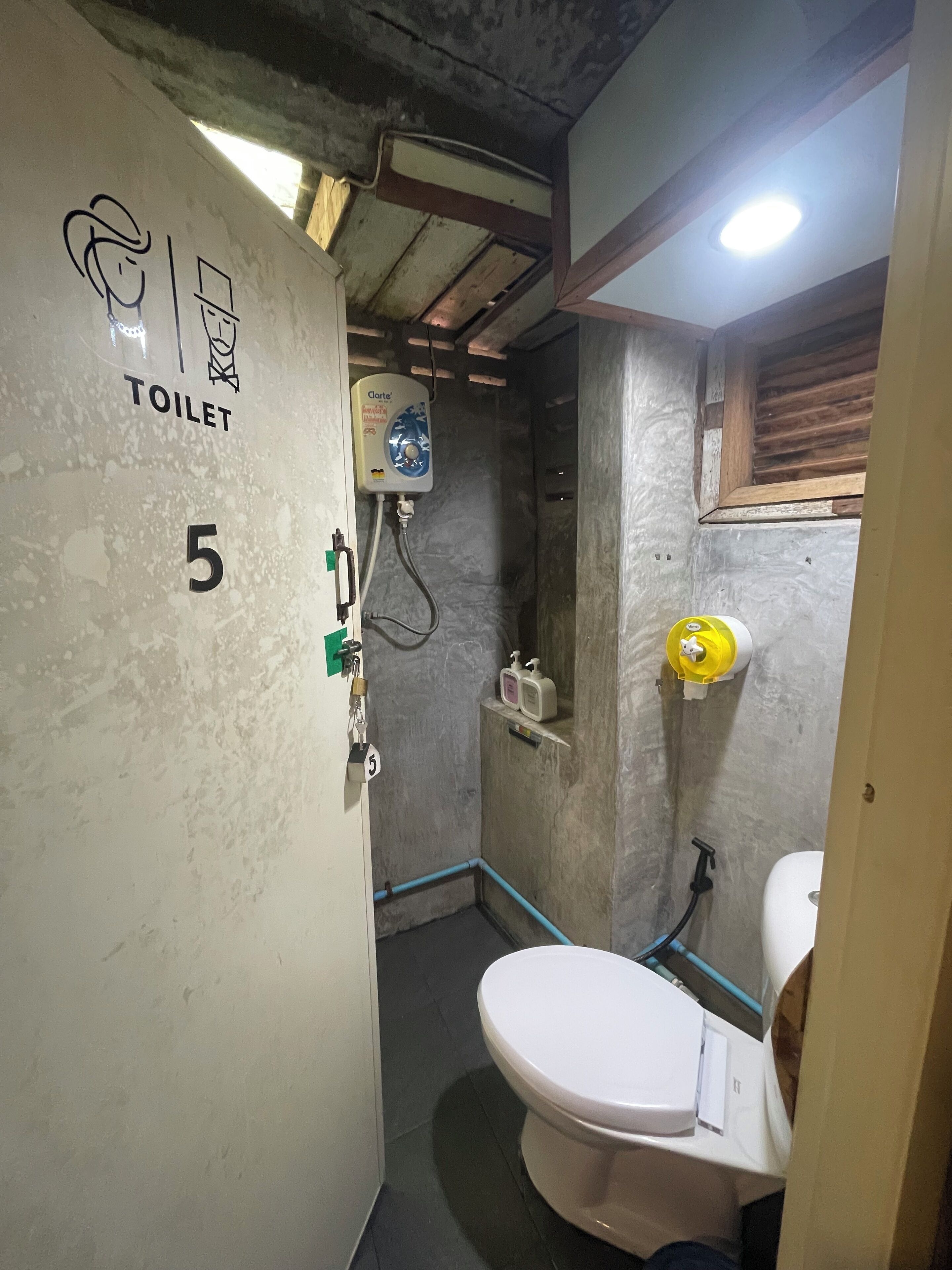 Une petite salle de bain compacte, équipée de toilettes, d’une douche et d’un sèche-mains. Les murs sont texturés et on peut y voir le chiffre « 5 ».