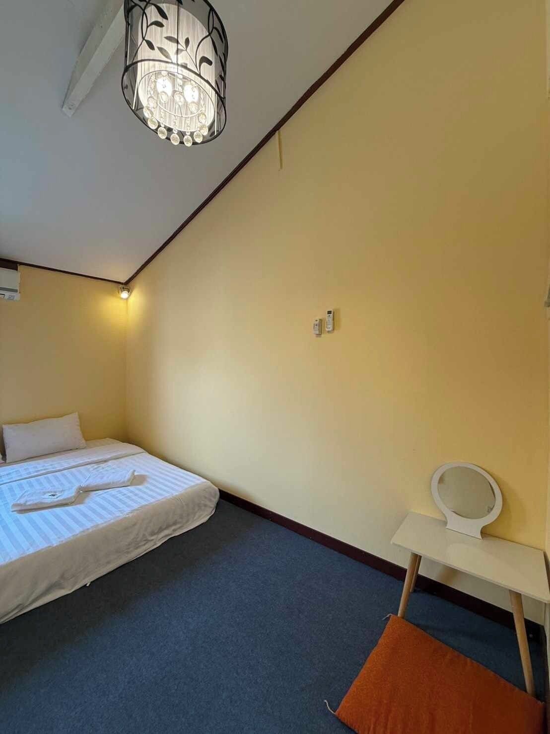 A small, well-lit room with a bed, a bedside table with a mirror, and a carpeted floor.