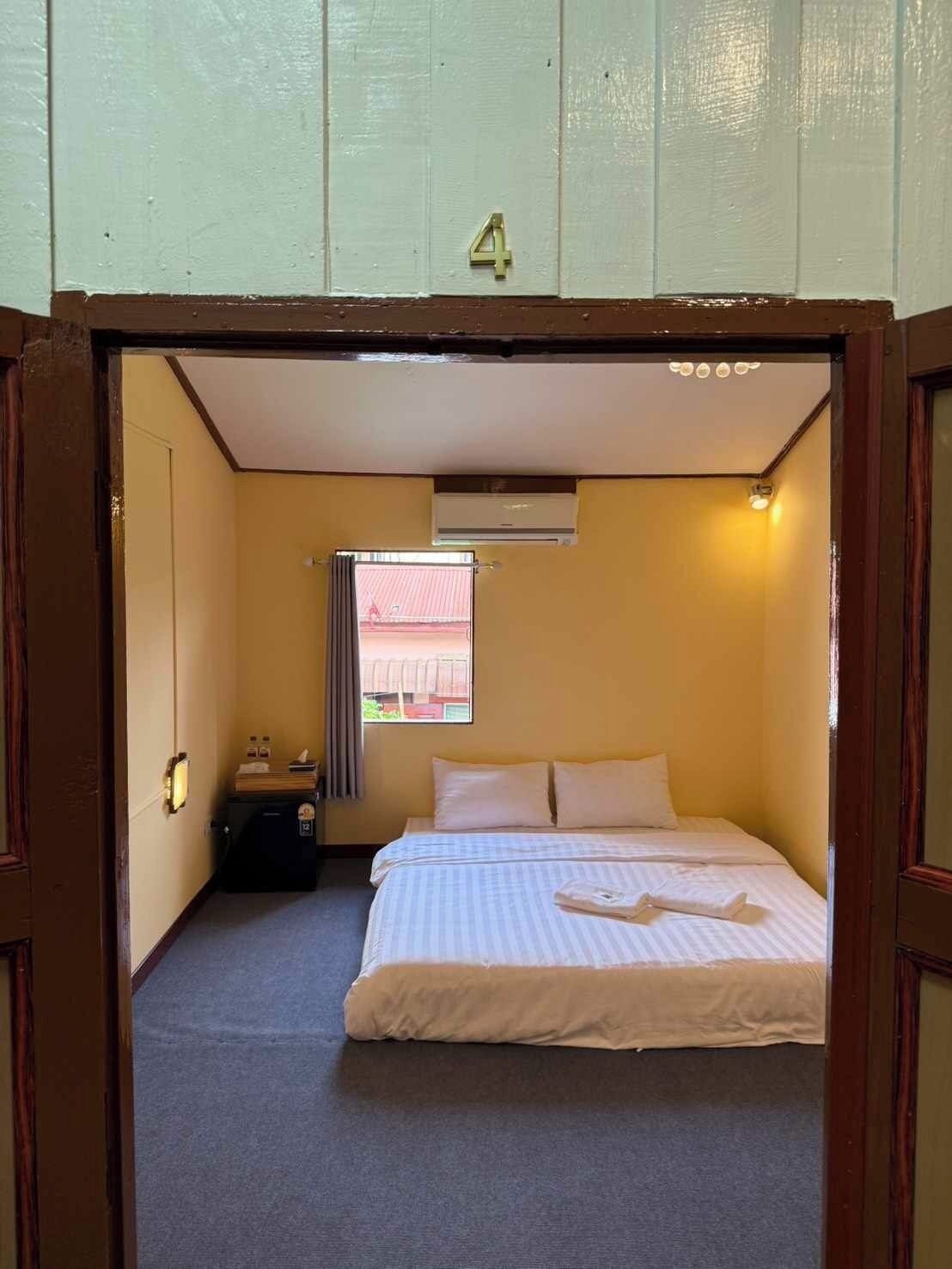A small, single-bed room with a window, a bedside table, and an air conditioning unit.