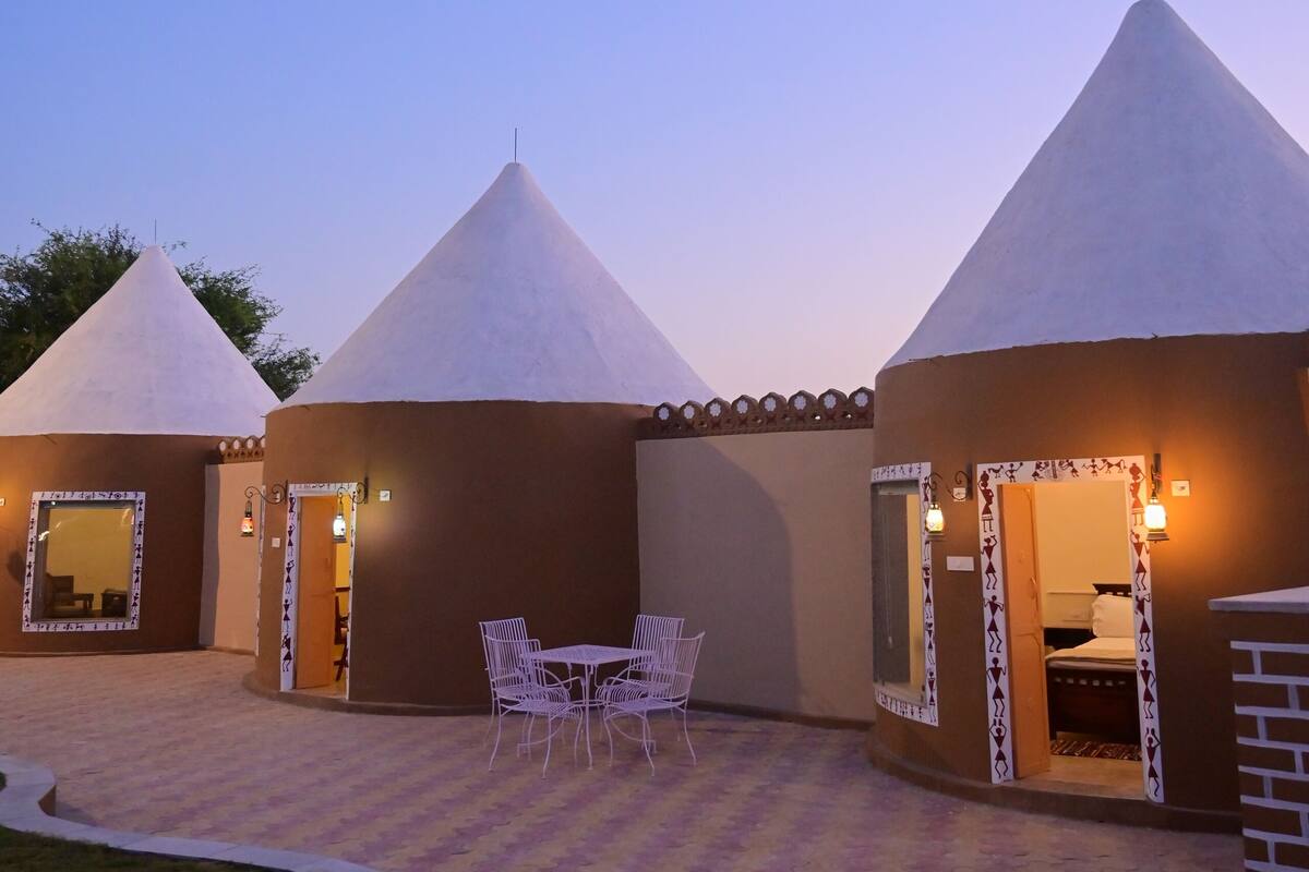 Un patio con tres construcciones de techo cónico, cada una con una puerta, una mesa pequeña y un sillón.