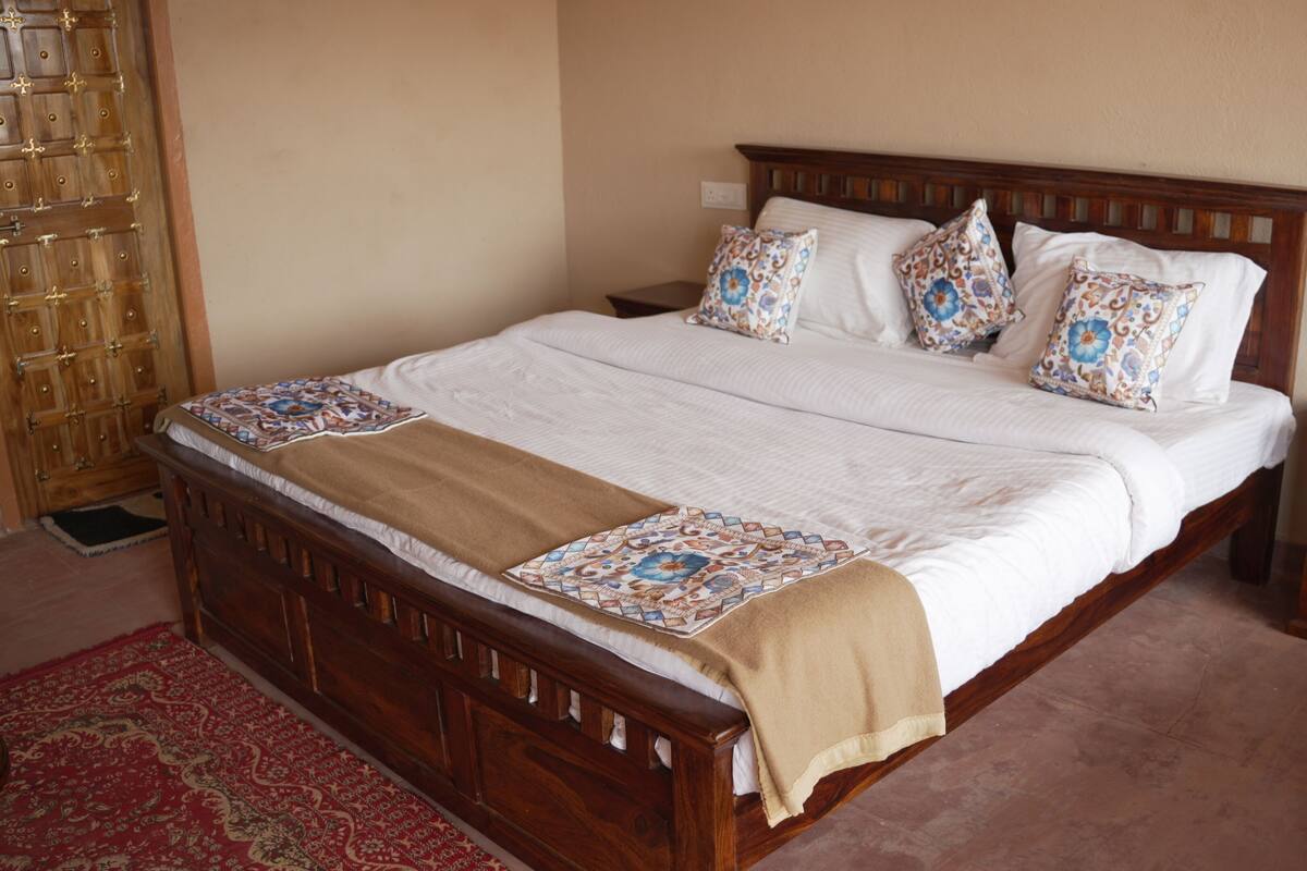 Una cama de madera con una colcha color beige y almohadas estampadas, una mesita de noche y un tapete rojo con diseño.
