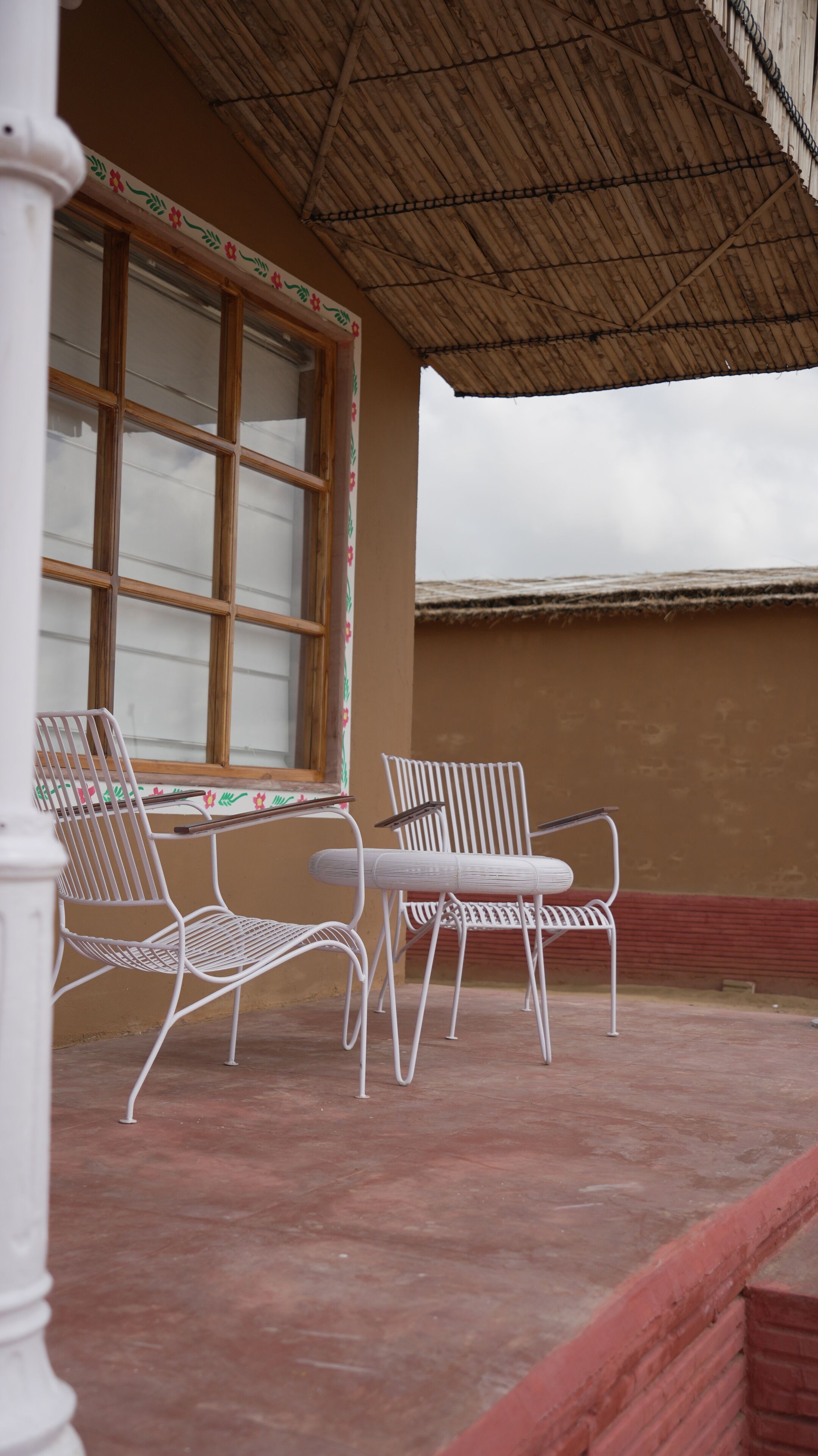 A covered patio area with two white metal chairs and a table, a large window with decorative trim, and a thatched roof.