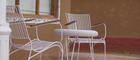 A covered patio area with two white metal chairs and a table, a large window with decorative trim, and a thatched roof.