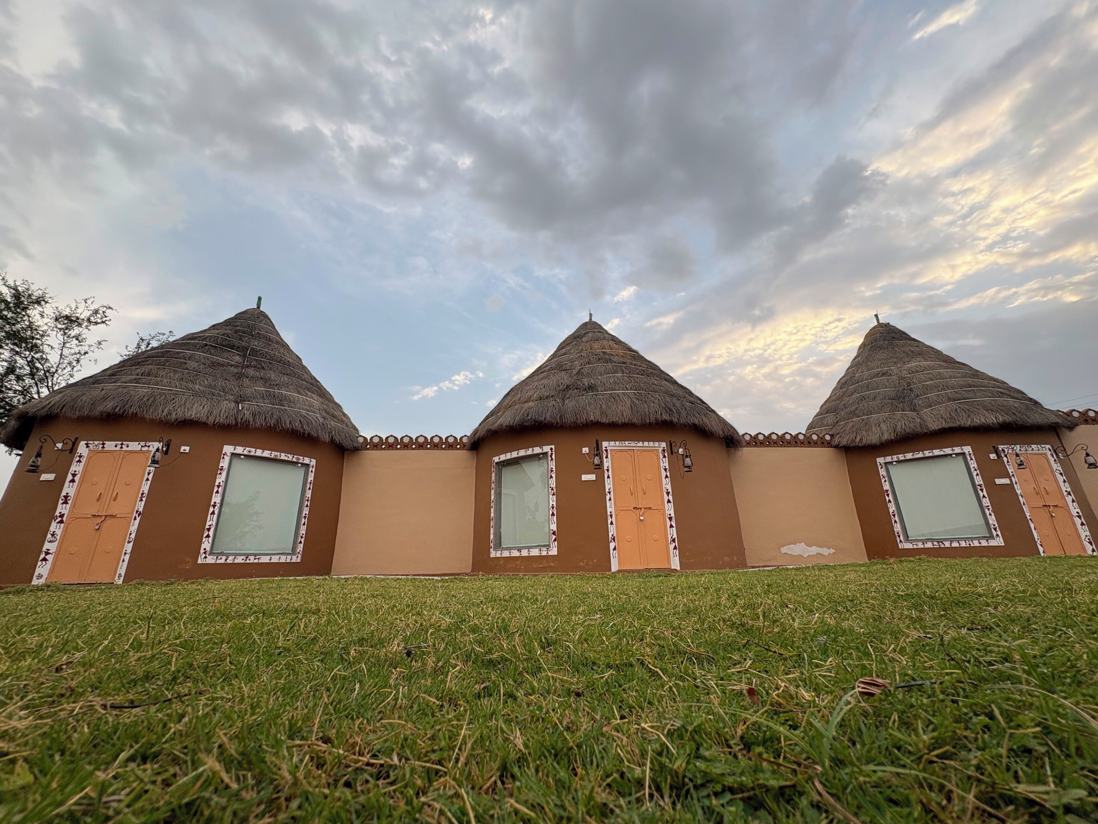 A row of three thatched-roof huts with windows and doors, set on a grassy slope.