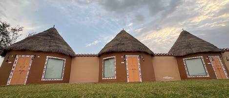 A row of three thatched-roof huts with windows and doors, set on a grassy slope.