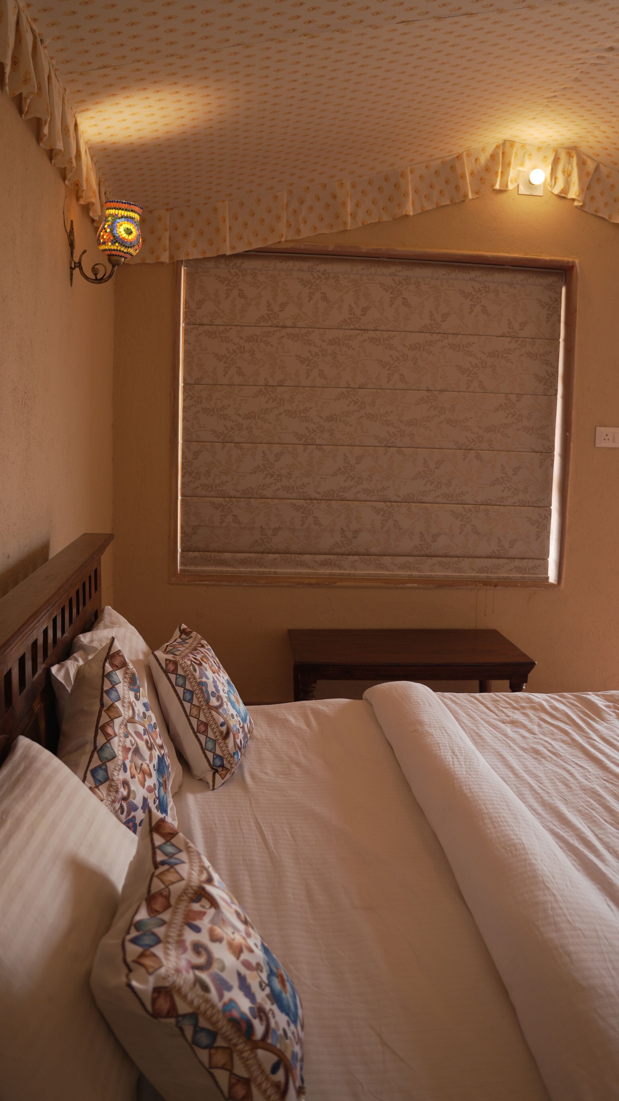 Een slaapkamer met een groot bed, een raam met bloemeninrichting, een wandlamp en een houten hoofdbord.