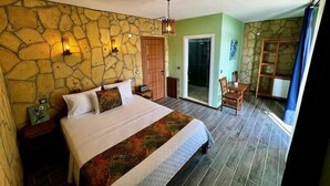 A bedroom with a stone wall, a bed with a patterned bedspread, a wooden chair, a small table, and a door leading to a balcony. - İda Lythos (Ayvacik)