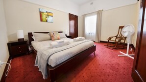 A hotel room with a large bed, a wooden nightstand, a chair, a fan, and a painting on the wall. - Beautifully restored 1930s gem, perfectly located in the heart of Portland. (Glenelg)