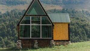 A cabin with a triangular roof, large windows, and a stone foundation, set against a forested hillside. - OKON Highlands Cottages & Camp (Yenokavan)